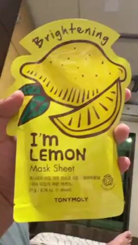 A hand holds up a yellow TonyMoly "I'm Lemon" mask sheet. The mask sheet is adorned with a cartoon drawing of a lemon. The words "Brightening," "I'm LEMON Mask Sheet," and the brand "TONYMOLY" are visible on the packaging. The camera zooms in on the mask sheet. A foot can be seen at the bottom of the frame. The background includes a wall and a window.