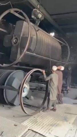 In what appears to be a manufacturing facility or industrial setting, four men, all wearing traditional Pakistani clothing including caps, are working around a large, cylindrical, black industrial machine. The machine is supported by large metal rings and has a motor attached to its top. The men are manipulating the metal rings, attempting to position or adjust them around the machine. The lighting in the facility is dim, but the general layout indicates a high-ceilinged industrial space. The floor is concrete.
