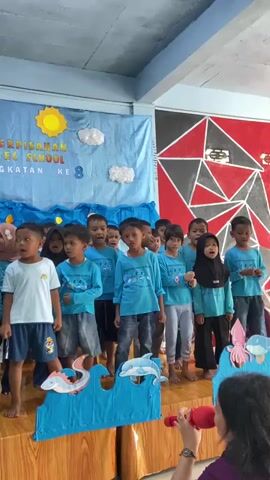 A group of young children in blue shirts stand on a stage decorated with a tropical theme. The backdrop features a palm tree, a sun, clouds, and the ocean. A banner behind the children reads "PERPISAHAN TK SCHOOL ANGKATAN KE 8". Several children are holding a microphone and singing. In front of the stage are cardboard cutouts of sea creatures like a jellyfish and dolphins. A woman is visible at the bottom right of the frame, holding a microphone. The children appear to be participating in a school event, possibly a graduation or farewell ceremony.