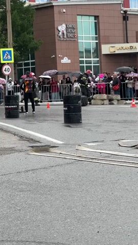 A group of firefighters in uniform are participating in a competition or demonstration on a paved street. They are wearing helmets and protective gear. The scene is outdoors, and there are spectators behind a metal fence. Some of the spectators are holding umbrellas, indicating it might be raining or drizzling. There are several black barrels and some concrete blocks set up as obstacles on the street. A pedestrian crossing sign is visible in the background. The overall atmosphere appears to be of an organized event.
