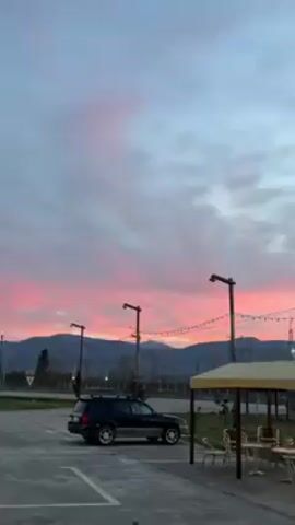 The video shows a parking lot with a black SUV parked on the left side. Two tall lampposts are visible with string lights hanging between them. Beyond the parking lot, there is a green field with trees, and in the background, there are mountains. The sky is predominantly gray with pink streaks from the sunset. The camera slowly pans to the right, revealing more of the field and mountain range.