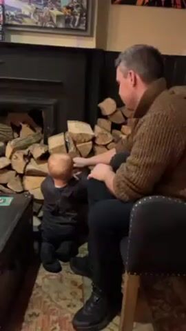 A man and toddler are sitting next to a pile of firewood. The man is seated in a dark-colored chair. He has grey hair, a brown knitted sweater, dark pants, and dark shoes. The toddler is dressed in dark clothing and is sitting on the floor reaching for a piece of wood. They are next to a black fireplace. The wood is stacked next to the fireplace. A dark trunk is on the floor to the left. The wall is dark except for framed artwork above the fireplace. The floor has a patterned rug.
