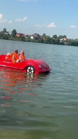 In this medium shot, three people are enjoying a sunny day on a calm lake in Dal'nie Prudishchi, Russia. A bright red pedal boat, designed to resemble a vintage car, glides across the water. Two adults and one child, all wearing bright orange life vests, are seated in the boat. The man in the front appears to be steering, while the woman behind him is looking towards the child. The child is seated in the front, facing forward.

The water is a clear, murky green, reflecting the blue sky and white clouds above. The shoreline in the background is lined with lush green trees and dotted with houses, suggesting a peaceful, rural setting. The time of day is clearly daytime, with bright sunlight casting shadows and illuminating the scene. The overall atmosphere seems relaxed and joyful, as the individuals enjoy a leisurely outing on the water. There is no visible text in the media file.
