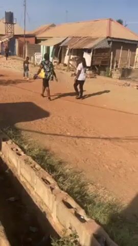 The video depicts a sunlit outdoor scene in what appears to be a developing or rural area. The ground is mostly dirt with some gravel, and there's a concrete drainage ditch running alongside the road in the foreground. Several small, modest buildings with corrugated metal roofs are visible in the background. A group of three individuals can be seen walking along the road; one is wearing a colorful patterned shirt, another is in a white t-shirt. The camera appears to be held low, and its movement suggests it's being carried while the videographer is walking. The shadow of a utility pole and the videographer is visible on the ground. The overall impression is a candid shot of a everyday life in a simple, somewhat impoverished setting.