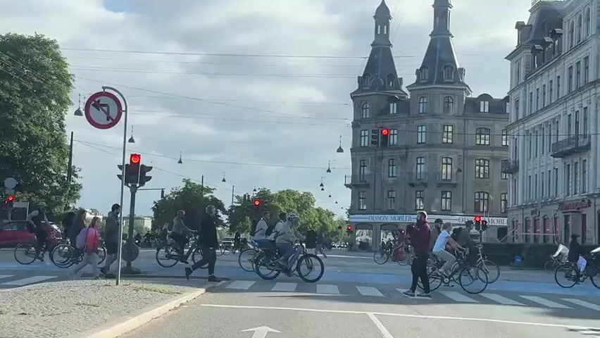The video shows a busy urban intersection with a pedestrian crossing. Traffic lights are red for vehicles, and a group of pedestrians and cyclists are crossing the street. People are walking and riding bicycles. A young girl with a pink backpack is walking across the crosswalk. Several cyclists are also crossing the street. In the background, there is a large building with distinct architecture, and on the right side of the street, there are more buildings and shops. The sky is overcast with clouds.