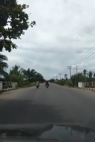 A wide-angle, first-person perspective shot shows a road with power lines running along the right side. The sky is overcast with gray clouds. On the road, there are two motorbikes, each with a rider. The motorbike in front is red, and the one behind it is white with a patterned rider. Palm trees and other vegetation line the left side of the road, with fields visible in the distance. The video appears to be shot from inside a vehicle, possibly a car, as indicated by the bottom edge of the frame showing a dark dashboard or hood. The motorbikes are moving away from the camera down the road.
