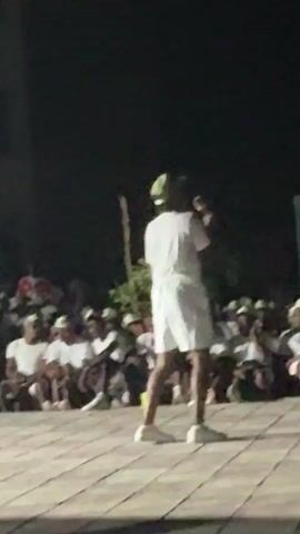 The video shows a person standing on an elevated brick platform, holding a microphone and speaking to a crowd of seated people. The speaker is wearing a white t-shirt, white shorts, white sneakers, and a light green hat. The crowd is primarily composed of young people, many of whom are wearing similar light green hats and white t-shirts. The setting appears to be outdoors at night, with a dark background. There are also some sparse bushes and a building visible. The speaker is gesturing with their hands as they address the crowd.