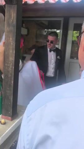 A wedding scene shows a bride in a white gown and a groom in a black tuxedo. The bride has bright red hair and a veil. She holds her dress with one hand while the groom holds a red ribbon attached to her dress. They are standing on a porch with a wooden pillar on the left. Another woman is visible in the background, and a young boy stands on the right, also holding a red ribbon. The groom kisses the bride on the cheek, and then she smiles. A small chocolate candy is on the ledge of the porch.