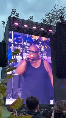 The video shows a large outdoor music festival stage with a massive LED screen. On the screen, a Black man wearing a black tank top, sunglasses, and a watch is singing into a microphone. He has tattoos on his arms and chest. Behind him, the screen displays images of musical instruments, likely a drum kit. The stage is adorned with pink and blue lights. To the right of the screen, large black speaker towers are visible. In the foreground, the backs of the heads of a crowd of people are visible, looking up at the stage. One person in the crowd is holding up a phone to record. The sky above is overcast and cloudy. The screen briefly changes to show another performer playing a saxophone, then shifts to display colorful lights and the word "PAR" in large, illuminated letters at the top right of the screen.
