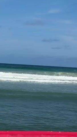 The video shows a view of the ocean from a beach. The sky is blue with a few clouds. The ocean is a deep blue-green color and there are waves rolling in. The waves are white and foamy. In the foreground, there is a red object, possibly a barrier or railing, and a white object, possibly a wall or structure. There is a person surfing on one of the waves.