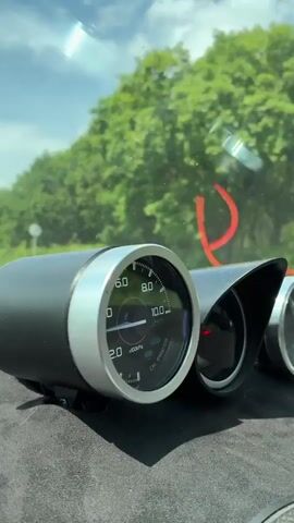 The video begins with a close-up of three gauges mounted on a car's dashboard. The leftmost gauge is an oil pressure gauge, displaying a needle that hovers around 6.0. The middle gauge is partially visible and angled away from the camera. The rightmost gauge is also partially visible. The background shows a blurred view through the windshield, with green trees and a bright sky. The camera then pans out and moves forward, showing the road ahead. A white semi-truck is visible in the distance, driving on a two-lane highway. Trees line both sides of the road, and the sky is blue with scattered clouds. The dashboard with the gauges is still visible in the foreground. The video ends as the camera continues to move forward, with the road and surrounding scenery filling the frame.
