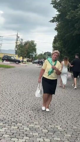 The video shows a woman in a yellow shirt and black skirt walking on a cobblestone path. She is carrying a white bag. In the background, several people are walking in the distance. There are cars and trees visible. The sky is overcast.