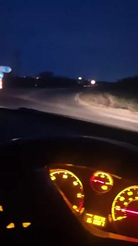 The view from inside a car driving on a dark, rural road at night. The dashboard is illuminated with yellow and red lights, and the speedometer shows the car is accelerating.  A stop sign is visible on the side of the road. The sky is dark blue.