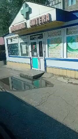 The video shows the exterior of a small pharmacy with a green cross symbol above the entrance. The pharmacy has large windows displaying various advertisements and products. In front of the pharmacy, there is a large puddle reflecting the building and the sky. The ground is paved with asphalt, and there is a raised brick path leading to the entrance of the pharmacy. The weather appears to be sunny.