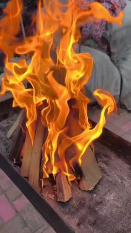 A close-up shot shows a small campfire burning in a metal tray. The fire is intense, with vibrant orange and yellow flames licking up from the stacked wooden logs. The logs are arranged in a teepee formation, with some appearing to be partially consumed by the fire. The tray is situated outdoors, with a paved surface visible beneath it, possibly a patio or courtyard. In the background, blurry objects suggest an outdoor setting, perhaps a backyard or campsite, with hints of furniture or stacked items. The flames are the primary focus, displaying dynamic movement and varying intensities.