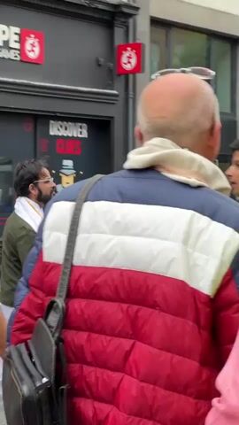 The video shows a back view of people walking down a street. In the foreground, a person wearing a puffer jacket with red, white, and blue stripes is visible. Behind them, another person is wearing a bright yellow safety vest with the text "ISKCON BELFAST" and a lotus flower logo. Other individuals are seen in the background, some wearing traditional Indian attire and others in casual clothing. The environment appears to be an urban setting with buildings and trees lining the street. The lighting suggests it is daytime, possibly overcast. The camera is positioned behind the first person, following them as they move.