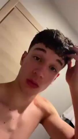 A shirtless young man with curly brown hair, fair skin, and prominent lips faces the camera, speaking. His right hand intermittently touches his hair. The background includes a light wood door and a white towel. He appears to be in a bathroom.