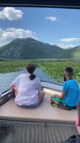 A boat moves through a calm waterway covered in lily pads, with a mountain range in the background under a cloudy sky. Two children are seated on the front of the boat, facing away from the camera, looking at the scenery. The child on the left is wearing a pink shirt and shorts, with their hair in a ponytail. The child on the right is wearing a blue shirt and green shorts, holding a tablet. The camera is positioned behind a third person whose head and shoulders are visible in the foreground. The boat is metallic and has upholstered seating. The water is dark and reflects the sky. The lily pads form a dense green carpet on the surface of the water, with a narrow channel of open water in the center. The mountain is covered in vegetation and rocky outcrops. The sky is blue with scattered white clouds.