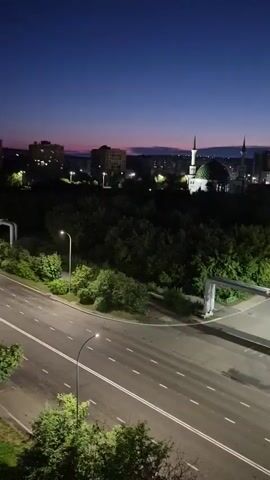 A wide shot captures an urban landscape at dusk or dawn. A dark blue sky with a faint pink and orange hue near the horizon forms the backdrop. In the middle ground, a lush green forest separates the foreground from the cityscape. A mosque with a green dome and two slender minarets stands out among the buildings. To the right of the mosque, modern buildings with illuminated windows and signs are visible. In the foreground, a paved road with white lane markings stretches across the frame, empty except for a single white truck driving away from the camera on the right side of the road. A large, arched pipe structure crosses the road from the right side. Streetlights illuminate sections of the road. Trees and vegetation line the sides of the road and the edge of the forest. The overall scene is quiet and still, suggesting early morning or late evening.