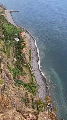 The video pans down from a cliff edge overlooking a coastline. The sea is a deep blue with white waves crashing onto a grey pebble beach. Along the beach, there are several small, orange-roofed buildings clustered together. Above the beach, the land is terraced and covered in greenery, with some areas appearing to be agricultural fields. A dirt path winds its way down the hillside. A narrow pier extends into the sea from the far end of the beach. The camera moves slowly, revealing more of the landscape. There are no people visible in the video.