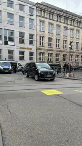 The video shows a pair of black Mercedes-Benz vans parked on a city street. One is parked and the other is in motion, making a left turn across yellow stripes painted on the road. A police motorcycle and car are visible nearby, suggesting a security detail. In the background is a Commerzbank building, indicating a commercial area. Pedestrians are standing near a barrier further down the street.