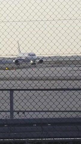 The video shows a wide shot of an airplane on a tarmac, viewed through a chain-link fence. The airplane is white with two engines visible under its wings. It is facing the camera and appears to be moving slowly. The tarmac is gray and has yellow and red lights on it. The sky is overcast. The fence is dark gray and has a diamond pattern.