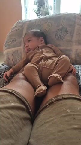 A close-up perspective shot shows a baby lying on someone's lap. The baby, wearing a tan onesie, is positioned with its back to the camera, head turned to the left. The person's legs, clad in tan pants, are prominent in the foreground. The baby is resting on a pillow that has a floral pattern. In the background, a window with blinds is visible.