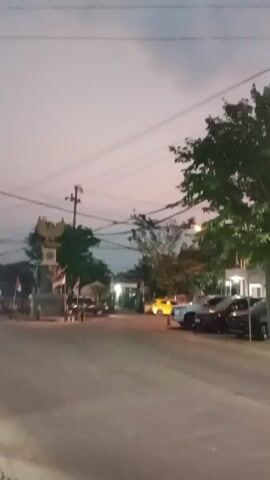 The video captures a street scene at dusk. A monument with a bird sculpture is visible on the left, adorned with flags. Several vehicles, including cars and motorcycles, are present on the road. People are riding motorcycles and walking. Power lines and street lights are visible overhead. The sky is a soft gradient of gray and pink, indicating the transition from day to night.