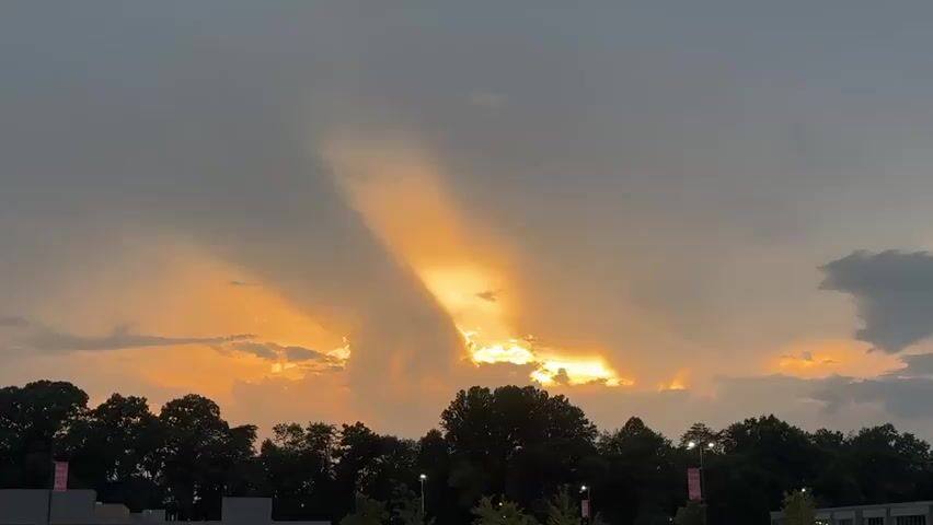 The video shows a sunset with crepuscular rays shining through the clouds. The sun is low on the horizon, casting a warm orange glow across the sky. The clouds are dark and ominous, but the sun's rays break through them, creating a dramatic effect. In the foreground, there is a line of trees and some buildings, which are silhouetted against the bright sky. The overall impression is one of natural beauty and power.