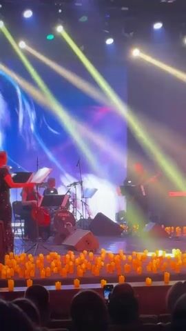 An indoor concert or musical performance is underway, featuring a band on a brightly lit stage. Numerous small, glowing yellow objects, resembling candles, are arranged across the front of the stage. The band consists of at least five musicians: a violinist on the left, a cellist, a drummer with a visible "TAMA" logo on their kit, an electric guitarist, and a keyboardist on the right. Large spotlights cast bright beams across the stage, which is also illuminated with blue and yellow stage lighting. A large screen behind the musicians displays dynamic, colorful abstract visuals. In the foreground, an audience is seated, with several individuals holding up mobile phones, seemingly recording the performance.