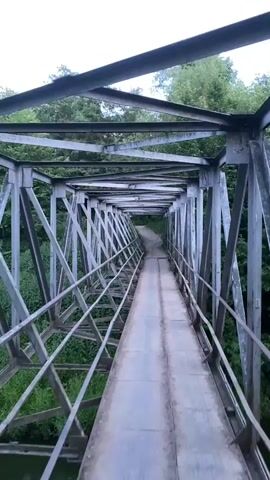 The video opens on a perspective of walking across a metal truss bridge. The bridge has a metal walkway and a metal railing on either side. Beyond the railing, trees and vegetation can be seen on both sides of a river. As the video progresses, the camera pans to the right, revealing a red brick building in the distance, surrounded by trees. The river below the bridge is visible, with water flowing rapidly. The camera continues to pan, showing more of the river and the surrounding trees. The video ends with a view of the river, its banks covered in greenery.