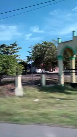 A vertical video shows a view from a moving vehicle. The scene is dominated by a clear blue sky with scattered clouds. On the left, an arched structure with pale yellow walls and green accents stands against a background of green foliage and trees. Further to the right, a white building with a black dome, resembling a mosque, is visible. The building has multiple arches and is surrounded by a grassy area and a low fence. The video captures a sense of passing by these structures, with motion blur indicating movement. The environment appears to be outdoors in a rural or semi-rural setting during daylight.