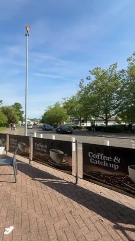 The camera pans from a roadside view in front of a sheltered cafe area to the parking lot beside it. The pavement is brick, and a banner advertising "Coffee & Catch Up" is visible along the roadside. Cars drive by on the road while the parking lot area shows parked vehicles, bike racks, and people walking through. The sky is clear and blue with sparse clouds. The lighting suggests it is daytime, possibly late morning or early afternoon.