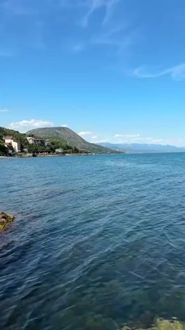 The media file captures a serene coastal scene under a bright, clear blue sky with scattered white clouds. The foreground is dominated by the calm, rippling turquoise waters of a sea or large lake. The water appears shallow in places, with glimpses of rocks and possibly underwater vegetation visible beneath the surface.

In the mid-ground, a gently sloping shoreline is visible, lined with buildings and vegetation. A road or pathway runs along the coast, with several houses and structures nestled amongst trees. A prominent hill or small mountain rises behind the settlement, its slopes covered in a mix of greenery and scattered buildings, including what appears to be a large, modern building or complex near the summit. The architecture suggests a resort or residential area. In the distance, a hazy mountain range can be seen on the horizon, underscoring the expansive natural landscape.

The scene is bathed in bright sunlight, indicating it is likely daytime, probably late morning or early afternoon, given the quality of light and the clear skies. The weather appears to be fair and pleasant, suitable for outdoor activities.

The location is identified as Bondarenkovo, Ukraine. While no people or specific objects are in sharp focus or prominently displayed, the overall impression is one of peaceful natural beauty and human habitation coexisting. The emotions evoked are those of tranquility, vastness, and perhaps a sense of vacation or relaxation. There is no visible text within the media file.