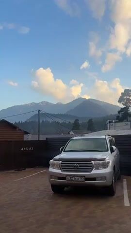 The video shows a stationary shot of a white Toyota Land Cruiser parked in front of a dark brown fence. Behind the fence, there are buildings and a utility pole with wires. In the background, there are lush green mountains covered in trees, with a hazy blue sky and some white clouds above. The overall scene appears to be a rural or mountainous landscape, possibly during the daytime due to the bright sky.