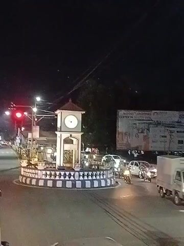 At night, a town square with a clock tower in the center is visible. The clock tower is white with a brown roof, surrounded by a decorative black-and-white curb and potted plants. Cars and motorcycles are circulating the square, illuminated by streetlights and headlights. A large billboard advertising a shopping center is located in the background. The traffic light is red.