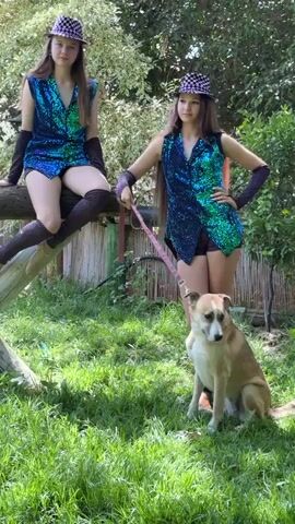Two young women, dressed in matching sequined vests and hats, pose with a dog in a grassy, outdoor area. The woman on the left is seated on a wooden beam, wearing thigh-high socks. The woman on the right stands and holds a leash attached to the dog, which is sitting in front of her. The environment appears to be a garden or park, with trees and foliage in the background.