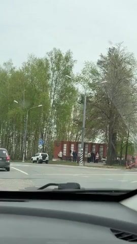 The video shows a driver's perspective from inside a car, approaching a small brick monument on the side of the road. The monument has a Soviet-era star emblem on it, and several people are standing around it. One person is walking towards the monument carrying flowers. There are birch trees lining the roadside, and a white SUV is parked nearby.  The weather appears overcast.  The car turns right at the intersection, passing the monument.  A lake can be seen through the trees behind the monument.