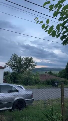 This static video captures a peaceful, overcast evening scene in Kosten, Bulgaria. The sky is filled with soft, grey clouds, and the diffused light suggests either dusk or early morning. In the foreground, lush green foliage hangs from the top right, partially out of focus. A chain-link fence with concrete posts runs horizontally across the mid-ground, separating the viewer from the main scene.

Behind the fence, a silver BMW E39 5-series sedan with black wheels is parked on the left, partially obscured by the fence. A portion of a white building is visible behind the car. A paved road stretches across the frame behind the fence. On the right side, a house with a reddish-brown tiled roof is nestled amongst trees and bushes, with a black wheelie bin visible near it.

Beyond the immediate structures, the landscape features numerous green trees and dense foliage, indicating a verdant environment. In the distant background, gentle rolling hills are visible under the cloudy sky, with hints of lighter, possibly pinkish, hues on the horizon, suggesting the last light of the day. Overhead, several utility lines traverse the top portion of the frame.

No people or animals are visible, and there is no discernible activity or movement. The scene evokes a quiet, serene, and somewhat contemplative atmosphere. No legible text is visible on any objects.