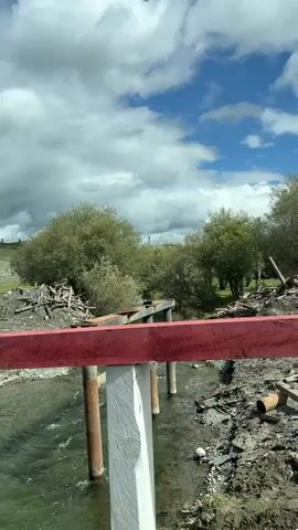 The video shows a view from a bridge overlooking a shallow, clear river. The bridge railing in the foreground is painted red and white. The riverbed is rocky, and debris, including logs and what appears to be building materials, is scattered along its banks. On the far bank, there are several trees and bushes. In the distance, more trees and hills are visible under a partly cloudy blue sky. The camera pans slightly, revealing more of the river and the surrounding terrain. The scene suggests a rural or natural environment, possibly undergoing some form of construction or repair.