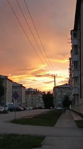 The video captures a city street scene during a vibrant sunset. Tall, multi-story buildings line the street on both sides, with balconies visible on some. Power lines stretch across the sky, intersecting the colorful clouds that dominate the frame. The sky is painted with hues of orange, yellow, and pink, indicating the sun is setting. On the street, several cars are parked and driving. A sidewalk runs along the right side of the street, and a grassy area is visible between the sidewalk and the road. Two people are walking on the sidewalk, one on the right and another further down the street. A bicycle can be seen in the distance on the road. The overall atmosphere is calm and serene, with the dramatic sky providing a beautiful backdrop to the urban environment.