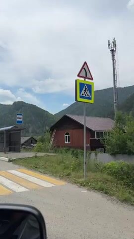 The video opens with a view of a bus stop in a rural setting, with a bus stop sign and shelter visible. A dirt road leads away from the bus stop, bordered by trees and houses. The camera pans to the right, revealing a red-painted house with a ladder leaning against it. The scenery then shifts to a wider view of rolling green hills and mountains under a partly cloudy sky. The road continues, with more houses and trees lining the sides. A white car is seen in the distance on the road. The video appears to be taken from inside a moving vehicle, as indicated by the reflections in the car's side mirror. The overall impression is one of a serene, natural landscape with signs of human habitation.