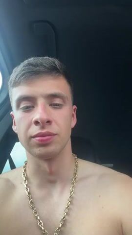 A young, shirtless man is seated in the driver's seat of a car, wearing a thick gold chain around his neck. He has short brown hair and light eyes, and he looks directly into the camera for a moment, then gestures with his right hand while the music plays in the background. The interior of the car is visible, with the dark ceiling and the rear seats in the background. The window is slightly open.