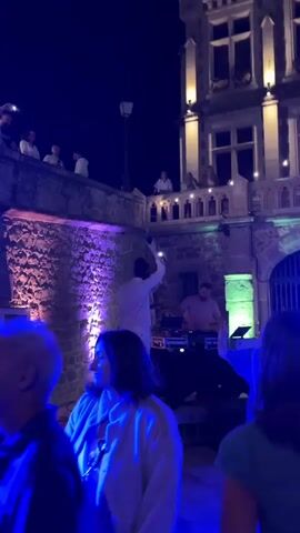 The video opens with a DJ in a white outfit standing on a platform behind a DJ booth, with a crowd of people watching and dancing in front of him. The scene is lit with blue and purple lights. As the video progresses, the camera pans across the crowd, showing people dancing and enjoying themselves. Some people are on a balcony overlooking the scene. The DJ is visible throughout the video, and appears to be interacting with the crowd. The video ends with a shot of the crowd dancing.
