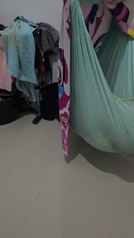 A close-up shot focuses on a light blue mesh hammock, decorated with colorful My Little Pony characters, hanging from a bar. The hammock is empty. To the left of the hammock, several items of clothing are draped over a clothes rack. The floor appears to be tiled.