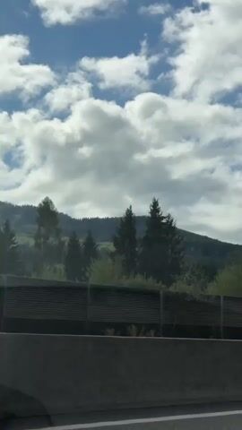 The video shows a view from a moving vehicle. In the foreground, a concrete barrier separates the road from the background. Beyond the barrier, there are trees and a hill covered in trees. A blue car is visible driving on the road in the second frame. Further in the background, there are houses and more trees. The sky is partly cloudy.