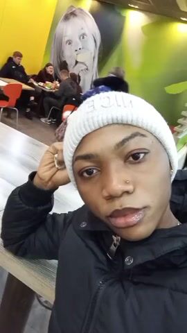 A close-up selfie shows a person with dark skin, wearing a white knit cap and a black puffy coat. They have a nose ring and are resting their head on their hand. They briefly purse their lips. In the background, other people are sitting at tables in what appears to be a fast-food restaurant. The wall is decorated with a yellow and green mural featuring a child eating. A man in a green jacket walks behind the subject. The lighting is bright and slightly yellow.