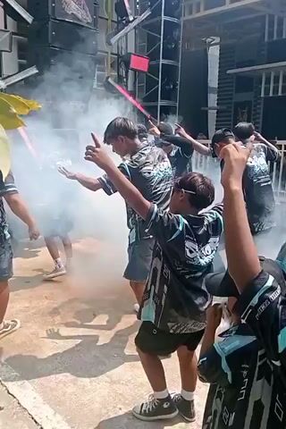 The video shows a group of people, many of whom are young boys, gathered outdoors. They are wearing matching dark shirts with a graphic design. There is a large sound system with speakers in the background, and smoke or fog is being emitted. Some individuals are dancing and gesturing with their arms. In the foreground, a woman is carrying a wooden plank on her head that holds several large cups filled with a dark liquid. Another woman in a white headscarf and patterned dress is standing to the right, looking towards the group. The environment appears to be a street or courtyard with buildings in the background. The overall scene suggests some kind of outdoor celebration or event.