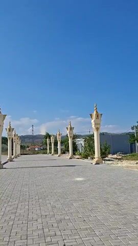 The video opens with a wide shot of a paved walkway lined with decorative pillars topped with golden finials. The pillars are illuminated by the sun against a clear blue sky. As the camera pans to the right, a stone wall with greenery along its base comes into view, followed by streetlights and trees. The scene then shifts to show large, white fabric canopies providing shade over a paved area. In the distance, a large, black, cube-shaped structure, possibly the Kaaba, is visible. The camera moves closer to the structure, revealing its black covering adorned with golden embellishments. More white canopies and decorative pillars are present around the structure. The overall environment appears to be an outdoor space, likely in a religious or ceremonial setting, under a bright, sunny sky with some scattered clouds. There are no people visible in the video.
