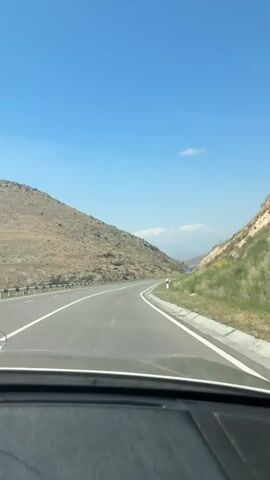 A vehicle is driving down a paved two-lane road, heading towards a mountain pass. The road is flanked by dry, grassy hills on both sides. The sky is clear and blue with a few scattered white clouds. The interior of the vehicle is visible in the foreground, including the dashboard and windshield. The video appears to be recorded from the perspective of the driver. There are no other vehicles or people visible in the immediate vicinity.